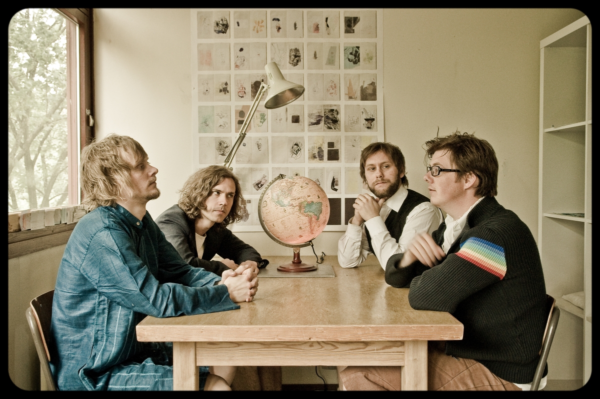 The band, shot from the side. Four young men looking at each other over a table